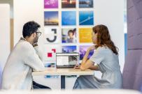 Two employees looking at a laptop seated in a collaboration space, with colourful branded wall pictures in the distance