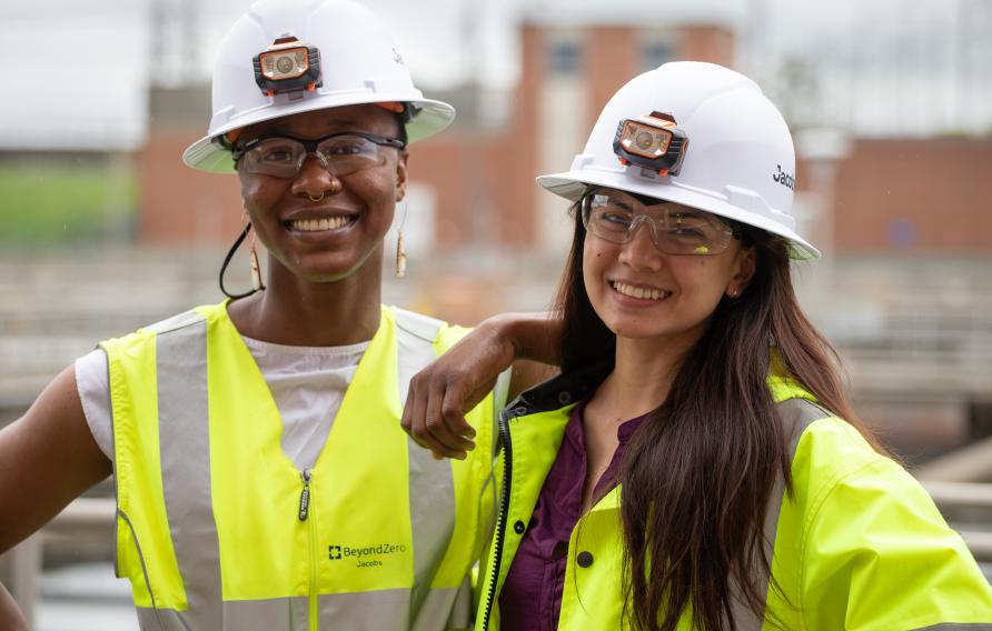 Two women in yellow PPE jackets and vests with white hard hats smile for the camera