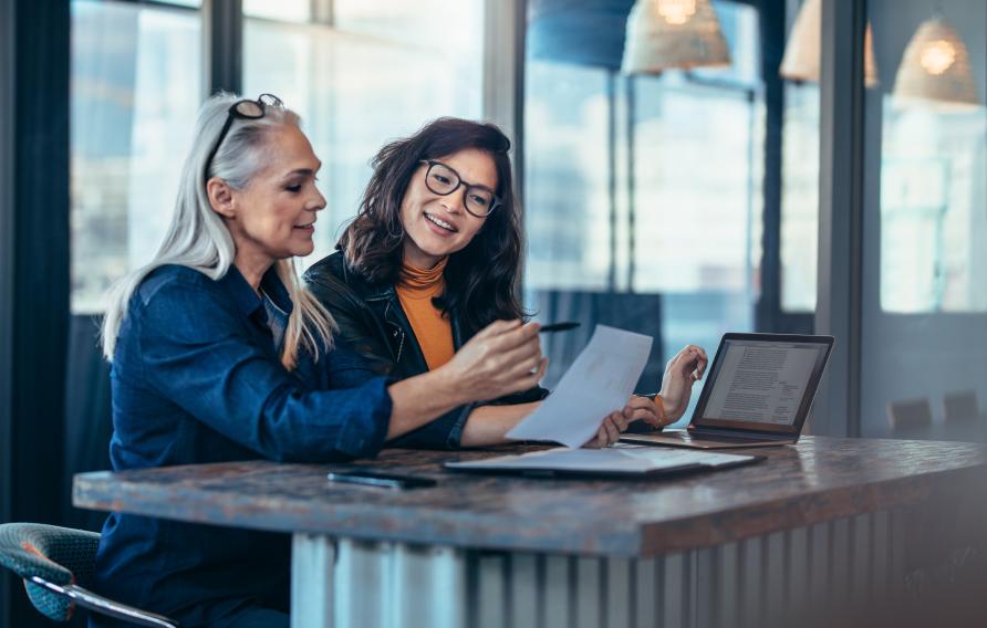 Two women analyzing documents while sitting on a table in office. Woman executives at work in office discussing some paperwork.