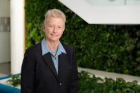 Blonde woman in a black suit and blue blouse in front of a greenery wall