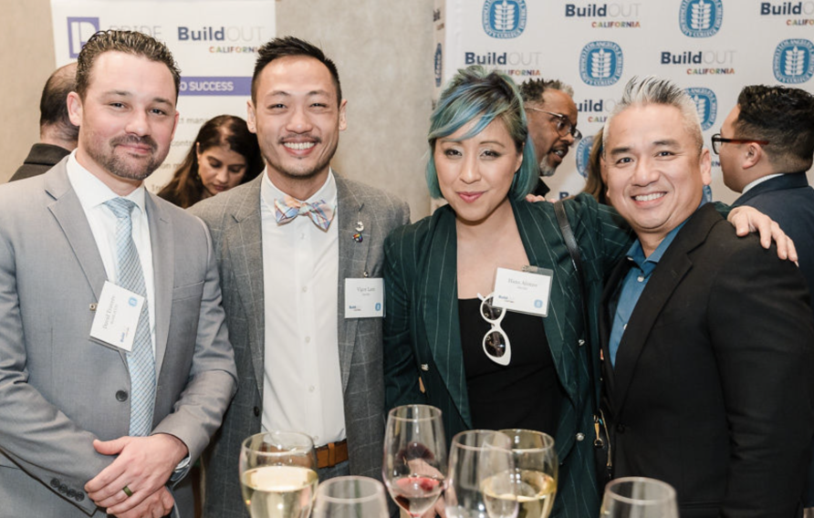 Group of men and women in business casual pose with wine goblets in front of a white and blue logo'd background